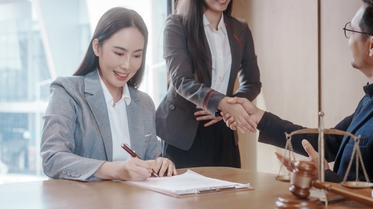 Close-up of businesspeople shaking hands across a desk with documents, pen, and a colleague standing in the background, suggesting a contract agreement or successful deal.